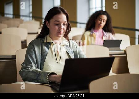 Kaukasische junge Erwachsene Frau mit Laptop, während sie im Hörsaal der Universität sitzt, polyethnische junge Erwachsene Frau im Hintergrund, die an einem Tablet arbeitet, beide konzentrierten sich auf akademische Aufgaben Stockfoto