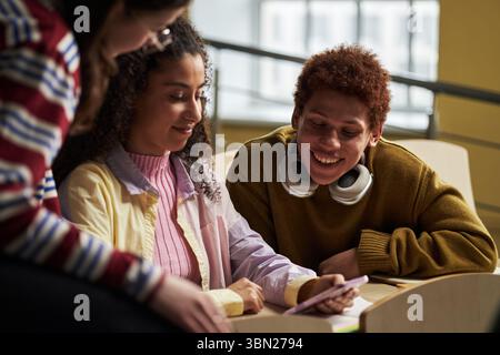 Gruppe multiethnischer Studenten, die zusammen sitzen, junge Frauen und Männer, die interagieren und lächeln, während sie sich das Smartphone ansehen, studieren oder an Projekten im Innenbereich zusammenarbeiten Stockfoto