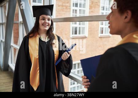 Kaukasische junge Frau, mit Mütze und Kleid, lächelnd, während sie die Diplommappe hält und mit einem anderen männlichen Mitschüler in akademischen Insignien im Universitätsgebäude spricht Stockfoto