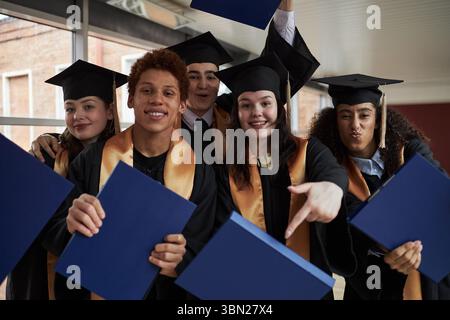 Gruppe multiethnischer Teenager, die Abschlussmützen und -Kleider tragen, um den Erfolg der Universität zu feiern, Diplommappen in der Hand zu halten, lächelnd und zusammen für die Kamera im Haus posiert Stockfoto