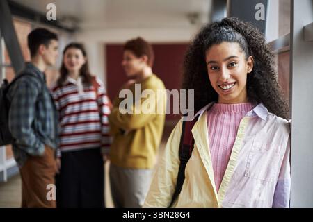 Porträt einer jungen Polyethnischen Schwarzen Frau, die in die Kamera lächelt, während sie im Flur der Universität steht und eine Gruppe junger Studenten im Hintergrund spricht Stockfoto