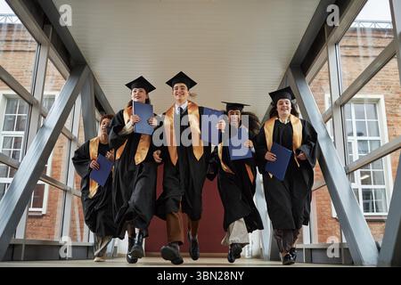 Gruppe multiethnischer junger Universitätsstudenten mit Abschlussmützen und -Kleidern, die zusammen auf der Campus-Brücke laufen, Diplome halten und nach der Abschlussfeier lächeln Stockfoto