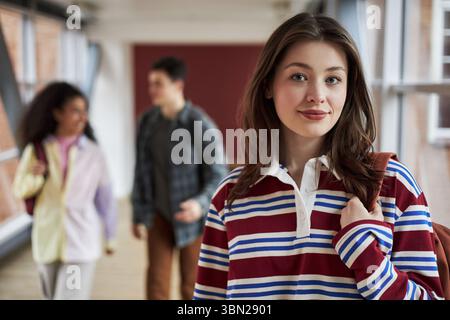 Porträt einer kaukasischen jungen Frau, die in die Kamera schaut, während sie im Flur des Colleges steht, mit einer multiethnischen Gruppe junger erwachsener Studenten, die im Hintergrund laufen Stockfoto