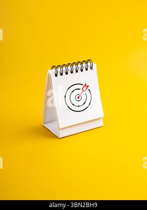 Zielpfeil-Bullseye-Symbol auf weißer Tischkalenderseite isoliert auf gelbem Hintergrund, vertikales Foto. Geschäftsziel und Erfolg, geplant, Agenda, Handeln Stockfoto