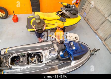 Zwei weibliche Mechaniker, die ein digitales Tablet und ein Multimeter verwenden, untersuchen den Motor eines Wasserfahrzeugs während der Wartung in der Werkstatt Stockfoto