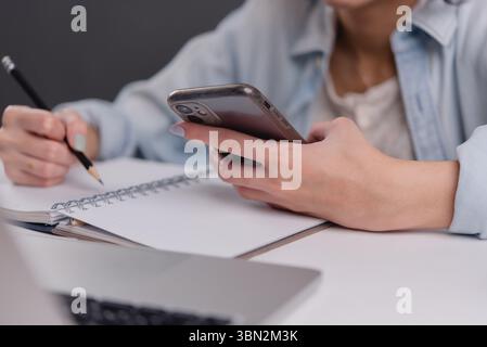 Junge Frau, die ein Smartphone benutzt, während sie Notizen macht und dabei die Balance zwischen digitaler Kommunikation und traditioneller Notiz in einem modernen Worksp zeigt Stockfoto