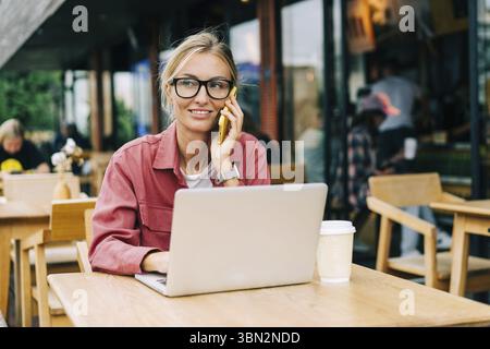 Junge attraktive kaukasische Frau, die in einem Café sitzt, arbeitet an einem Laptop und spricht am Telefon Stockfoto