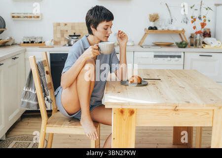 Eine Frau sitzt in der Küche, frühstückt und trinkt Kaffee Stockfoto
