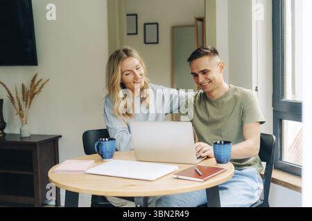 Junge glückliche, verliebte Paare sitzen zu Hause am Tisch, trinken Kaffee und arbeiten am Laptop Stockfoto