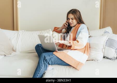 Eine junge Geschäftsfrau in einem Cardigan, die auf der Couch sitzt, arbeitet an einem Laptop und spricht am Telefon Stockfoto