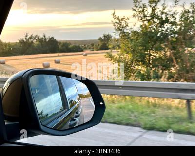 Ein Blick aus dem Seitenspiegel eines Autos auf der deutschen Autobahn, der die Landschaft neben der Straße bei Sonnenuntergang zeigt Stockfoto