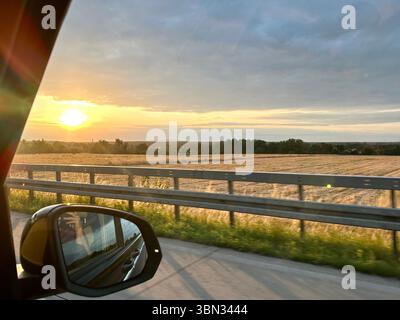 Ein Blick aus dem Seitenspiegel eines Autos auf der deutschen Autobahn, der die Landschaft neben der Straße bei Sonnenuntergang zeigt Stockfoto