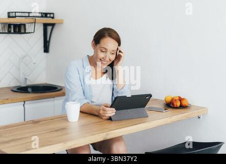 Junge, attraktive, glueckliche Frau, die in der Kueche sitzt und sich auf einem digitalen Tablet die sozialen Medien ansieht Stockfoto