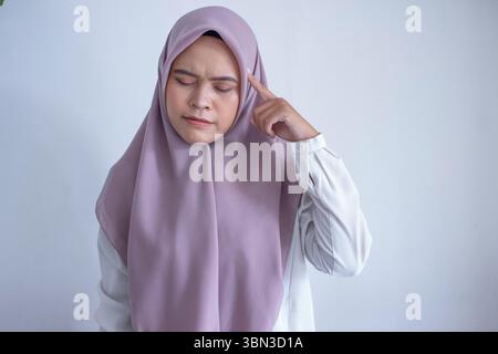 Die junge asiatische muslimische Frau, die einen violetten Hijab trägt, schaut mit dem Finger nach links. Sie ist eine denkende Frau Stockfoto