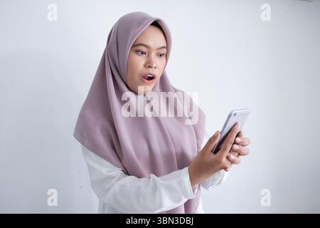 Die junge muslimische asiatin trägt einen violetten Hijab und hält ein Handy. Sie ist von etwas am Telefon überrascht oder schockiert Stockfoto