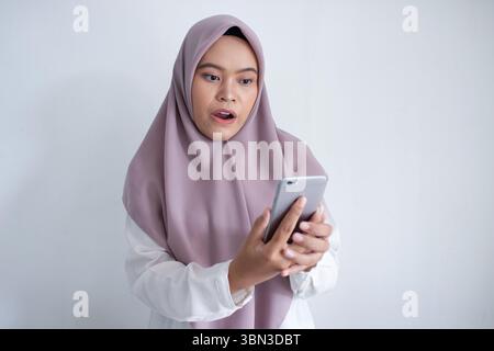 Die junge muslimische asiatin trägt einen violetten Hijab und hält ein Handy. Sie ist von etwas am Telefon überrascht oder schockiert Stockfoto