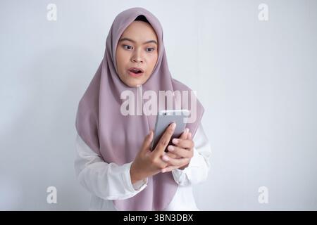 Die junge muslimische asiatin trägt einen violetten Hijab und hält ein Handy. Sie ist von etwas am Telefon überrascht oder schockiert Stockfoto