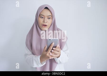 Die junge muslimische asiatin trägt einen violetten Hijab und hält ein Handy. Sie ist von etwas am Telefon überrascht oder schockiert Stockfoto