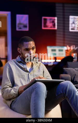 Ein junger, fokussierter Mann, der sich darauf vorbereitet, E-Mails zu beantworten und tägliche Aufgaben zu erledigen, arbeitet gern von seinem modernen Apartment aus. Person, die auf dem Laptop im Internet surft und sich auf dem Sofa entspannt. Stockfoto