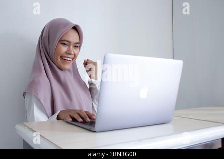 Die junge muslimische asiatische Frau, die einen violetten Hijab-Schal trägt, lächelt und hält ihre Hände hoch in der Luft, während sie auf einen Laptop blickt Stockfoto