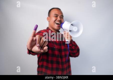 Energischer indonesischer Mann im Flanellhemd-Outfit, der Megaphon hält und schrie und stolz den kleinen Finger zeigt, der nach der Wahl in violette Tinte getaucht ist Stockfoto