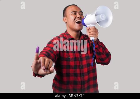 Energischer indonesischer Mann im Flanellhemd-Outfit, der Megaphon hält und schrie und stolz den kleinen Finger zeigt, der nach der Wahl in violette Tinte getaucht ist Stockfoto