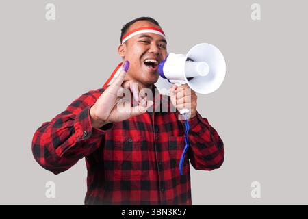 Energiegeladener indonesischer Mann im Flanell-Hemd-Outfit, Fahnenhaarband, der Megaphon hält und schrie und stolz den kleinen Finger zeigt, der in Purpur getaucht ist Stockfoto