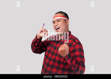 Energiegeladener indonesischer Mann in Flanellhemd-Outfit, Fahnenhaarband, der stolz den kleinen Finger zeigt, der nach der Wahl in violette Tinte getaucht ist Stockfoto