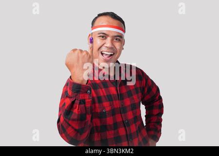 Energiegeladener indonesischer Mann in Flanellhemd-Outfit, Fahnenhaarband, der stolz den kleinen Finger zeigt, der nach der Wahl in violette Tinte getaucht ist Stockfoto