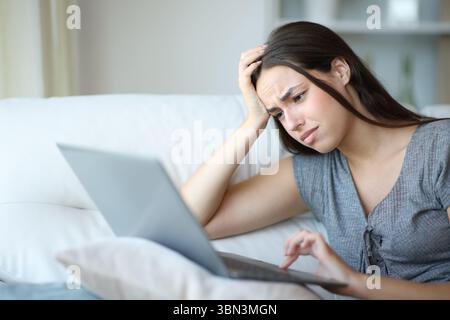 Eine gestresste Frau, die schlechte Nachrichten auf dem PC liest und zu Hause auf einer Couch sitzt Stockfoto