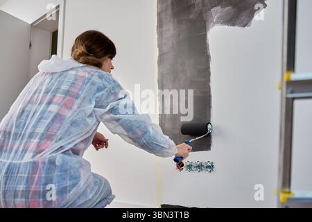 Frau in Schutzanzug Hose weiße Wand mit schwarzem Druck mit Rolle während der Renovierung. Weibliche Arbeiterin, die die Innenwand anstrich. Konzept des Home Impro Stockfoto