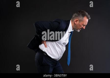 Reifer Mann in einem Anzug, der seinen unteren Rücken schmerzhaft hält, sich nach vorne beugt und grimmt, was auf schwere Lendenwirbelbeschwerden oder Rückenverletzungen hinweist. Stockfoto