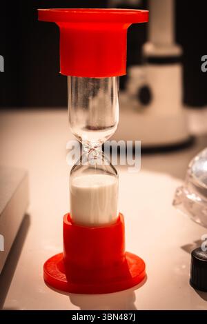 Kleine rote Sanduhr mit weißem Sand auf dem Tisch. Sandglas auf Labortisch. Zeitvertreib-Konzept. Zeitmessgeräte. Vintage-Uhr. Retro-Timer. Stockfoto