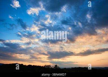 Atemberaubender Himmel voller dramatischer Wolken übergeht bei Sonnenuntergang von tiefem Blau zu sanften Pastelltönen. Horizont gesäumt von silbrigen Hügeln und Wäldern, die der ruhigen und weitläufigen Landschaft Tiefe verleihen. Stockfoto