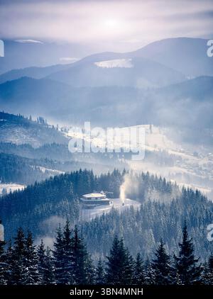 Winterliche Berglandschaft. Einsame Hütte inmitten eines dichten Waldes mit schneebedeckten Kiefern, mit Rauch aus dem Kamin. Sanfte Hügel, Täler mit Schnee. Weiches Sonnenlicht filtert durch Nebel. Stockfoto