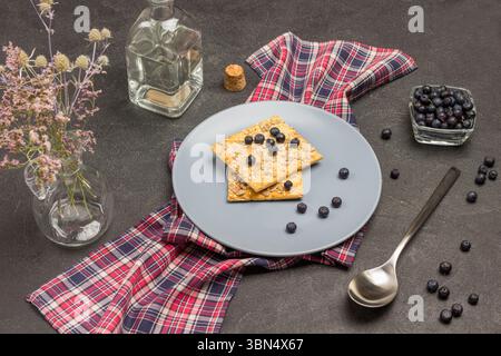 Grauer Teller mit Keksen und Heidelbeeren auf karierter Serviette. Mineralwasser. Heidelbeeren in Glasschale. Der Löffel liegt auf dem Tisch. Schwarzer Hintergrund. Top VI Stockfoto