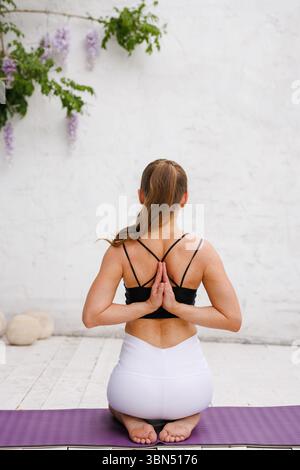 Frau im Yoga-Outfit sitzt im umgekehrten Gebet auf der Matte drinnen und konzentriert sich auf Rückenflexibilität und achtsame Wellness-Praxis Stockfoto
