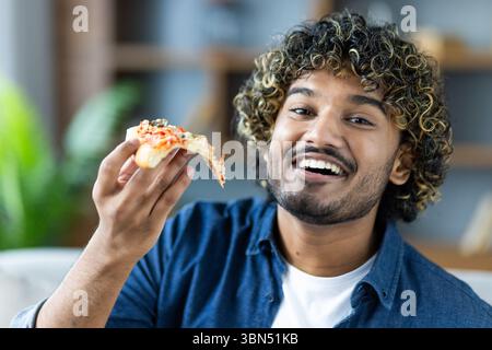Ein lächelnder Mann mit lockigen Haaren genießt ein Stück Pizza und fängt die Freude an einer einfachen Mahlzeit und einem glücklichen Moment ein. Stockfoto