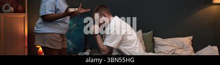 Schwarze Frau, die mit den Händen steht, während ein kaukasischer Mann auf dem Bett sitzt und sich mit der Hand auf dem Gesicht nach vorne beugt, während der Konflikt im Schlafzimmer Anzeichen von Bedrängnis zeigt Stockfoto