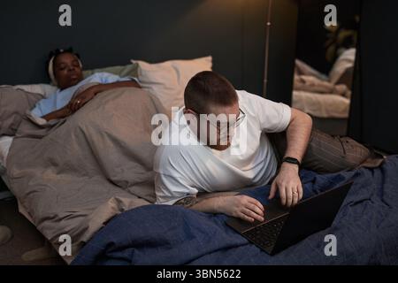 Ein kaukasischer Mann mittleren Alters benutzt Laptop, während er auf dem Bett neben der schwarzen Frau liegt, die unter der Decke ruht, wobei er Augenkontakt vermeidet und distanziert erscheint, was Beziehungskonflikte veranschaulicht Stockfoto
