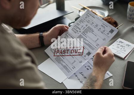 Kaukasischer junger erwachsener Mann mit überfälliger Rechnung, der am Tisch sitzt, Finanzdokumente und handschriftliche Notizen sichtbar, die Stress und Besorgnis während der Konfliktsituation zeigen Stockfoto