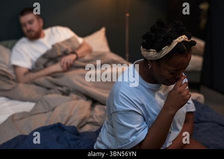Junge Erwachsene Schwarze Frau, die weinend auf dem Bett sitzt, junger Erwachsener kaukasischer Mann, der hinter ihr unter Decke sitzt und sie beobachtet, beide erscheinen aufgebracht während des Konflikts im Schlafzimmer Stockfoto