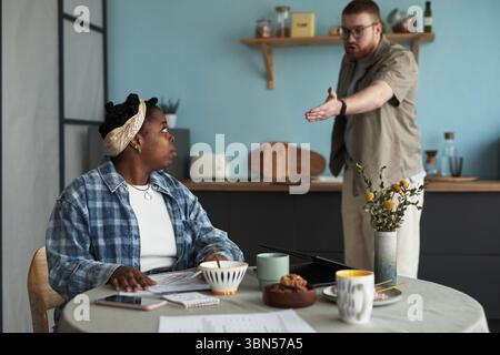 Schwarze junge Erwachsene Frau, die am Tisch sitzt und auf kaukasischen jungen erwachsenen Mann blickt, der während des Konflikts wütend steht und Gesten macht, Dokumente und Laptop auf dem Tisch, Küchenhintergrund sichtbar Stockfoto