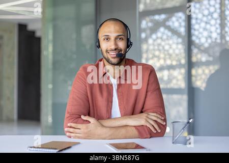 Ein lächelnder Kundendienstmitarbeiter, der ein Headset in einer modernen Büroumgebung trägt. Er sitzt an einem Schreibtisch. Stockfoto