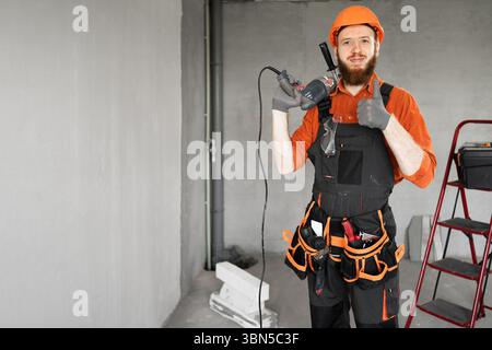 Porträt eines bärtigen Reparaturarbeiters mit orangefarbenem Helm und Overall, der den Daumen nach oben zeigt und den Bohrlochbohrer auf der Baustelle hält. Kopierbereich Stockfoto