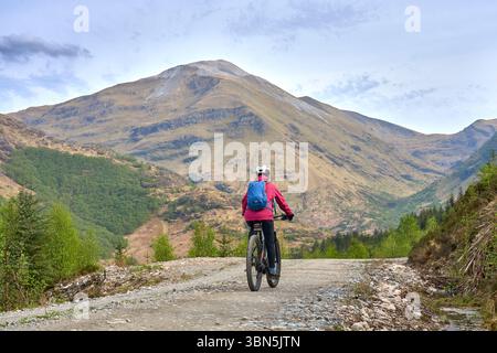 Aktive Seniorin, die ihr E-Mountainbike im Glen Nevis Tal unterhalb des Ben Nevis Gipfels im Hochland nahe Fort William in Schottland fährt. Stockfoto