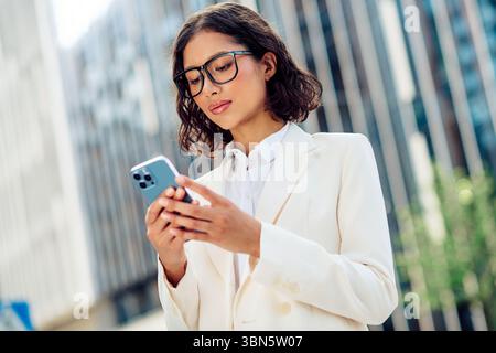 Selbstbewusste Geschäftsfrau, die ihr Smartphone im urbanen Umfeld nutzt, während sie draußen bei Tageslicht steht und Eleganz und Professionalität zeigt Stockfoto