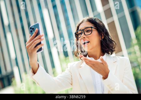 Selbstbewusste junge Geschäftsfrau, die ein Smartphone draußen vor städtischen Wolkenkratzern hält, während sie präsentiert oder kommuniziert. Stockfoto