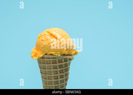 Ein einzelner Messlöffel Orangeneis steht auf einem Waffelkegel vor einem hellblauen Hintergrund. Stockfoto