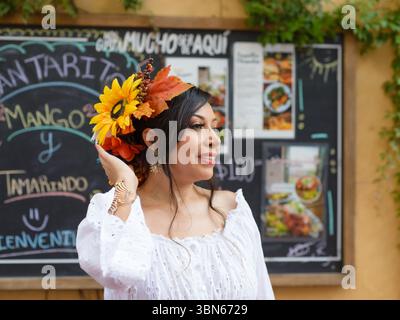 Eine Frau mit Sonnenblume und Herbstblatt lächelt, die vor einem Tafel-Menü steht. Sie trägt ein weißes Ösenoberteil und Goldschmuck. Stockfoto